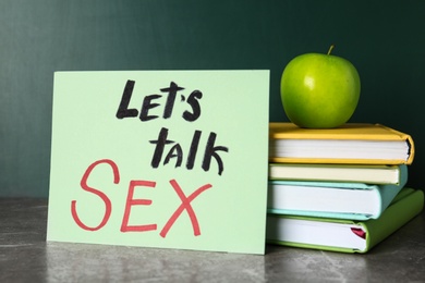 Photo of Books, apple and sign with phrase "Let's talk sex" on grey table near chalkboard