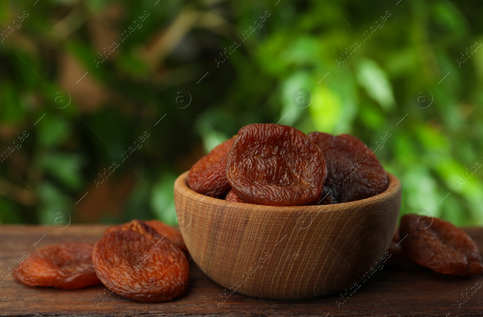 Photo of Bowl of tasty apricots on wooden table against blurred green background, space for text. Dried fruits
