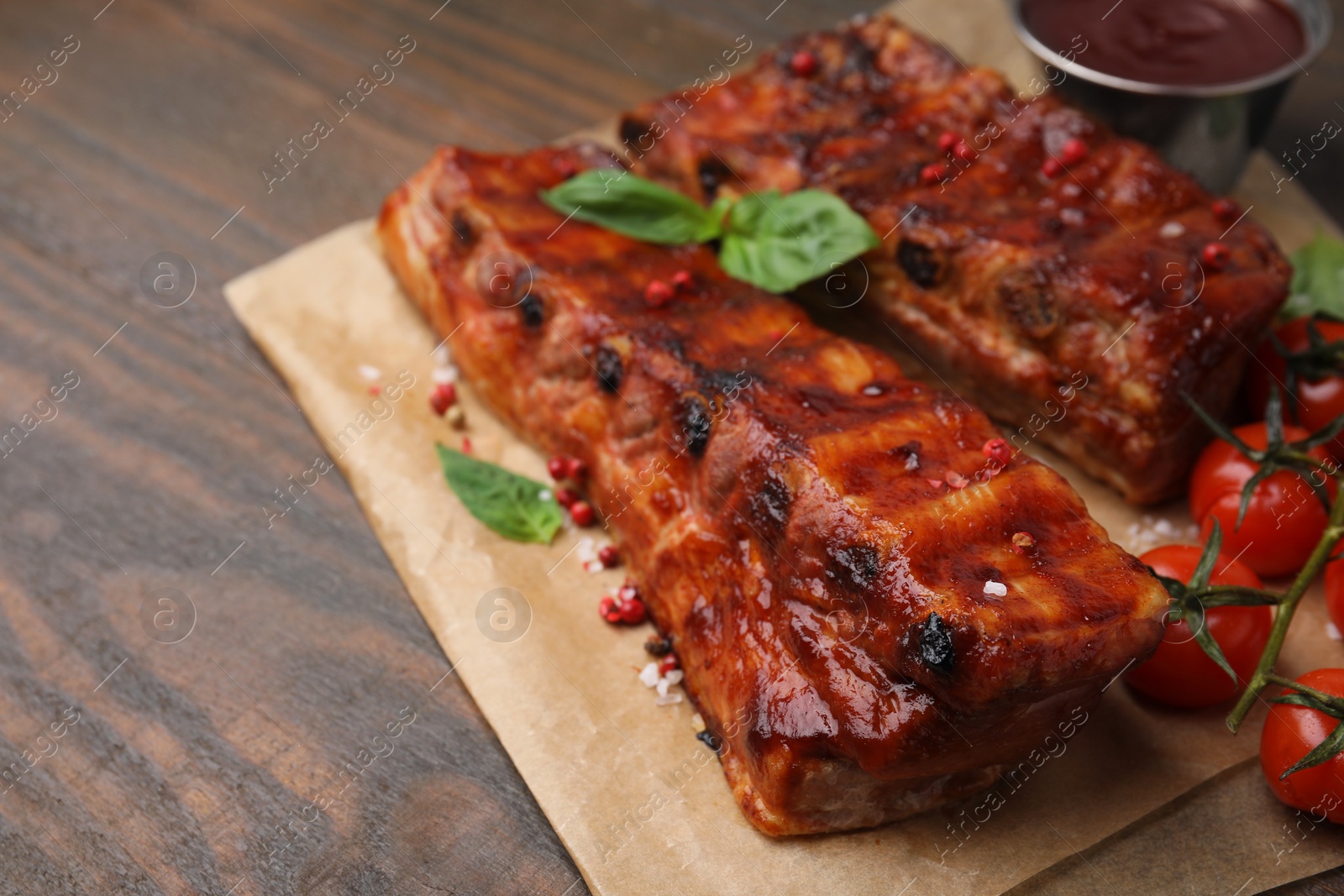 Photo of Tasty roasted pork ribs served with sauce, basil and tomatoes on wooden table, closeup. Space for text