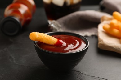 Photo of Tasty cheesy corn stick in bowl of red sauce on black table, closeup
