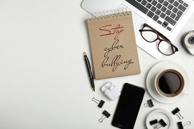 Notebook with phrase STOP CYBER BULLYING, laptop and cup of coffee on white table, flat lay. Space for text