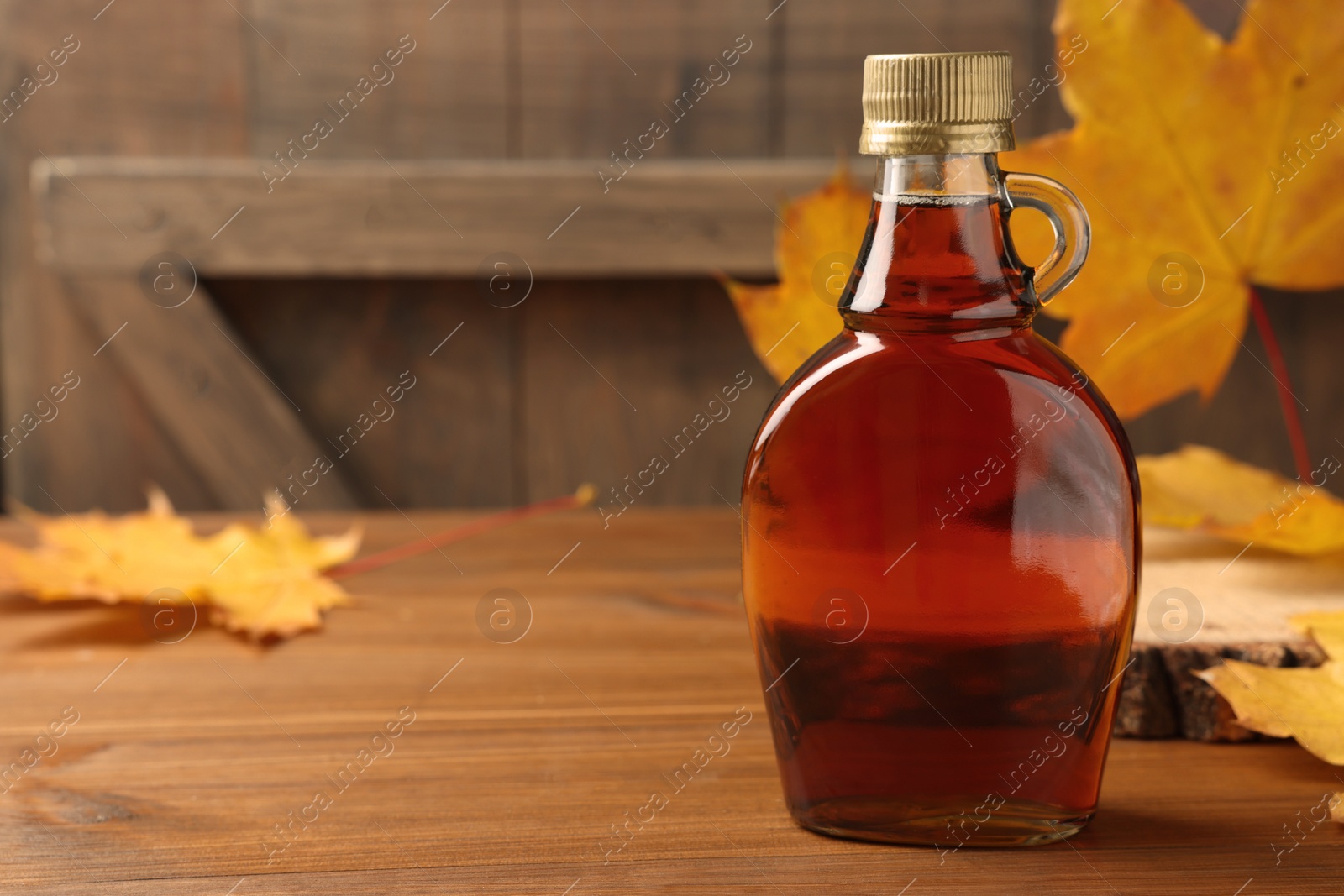 Photo of Bottle of tasty maple syrup and dry leaves on wooden table, space for text