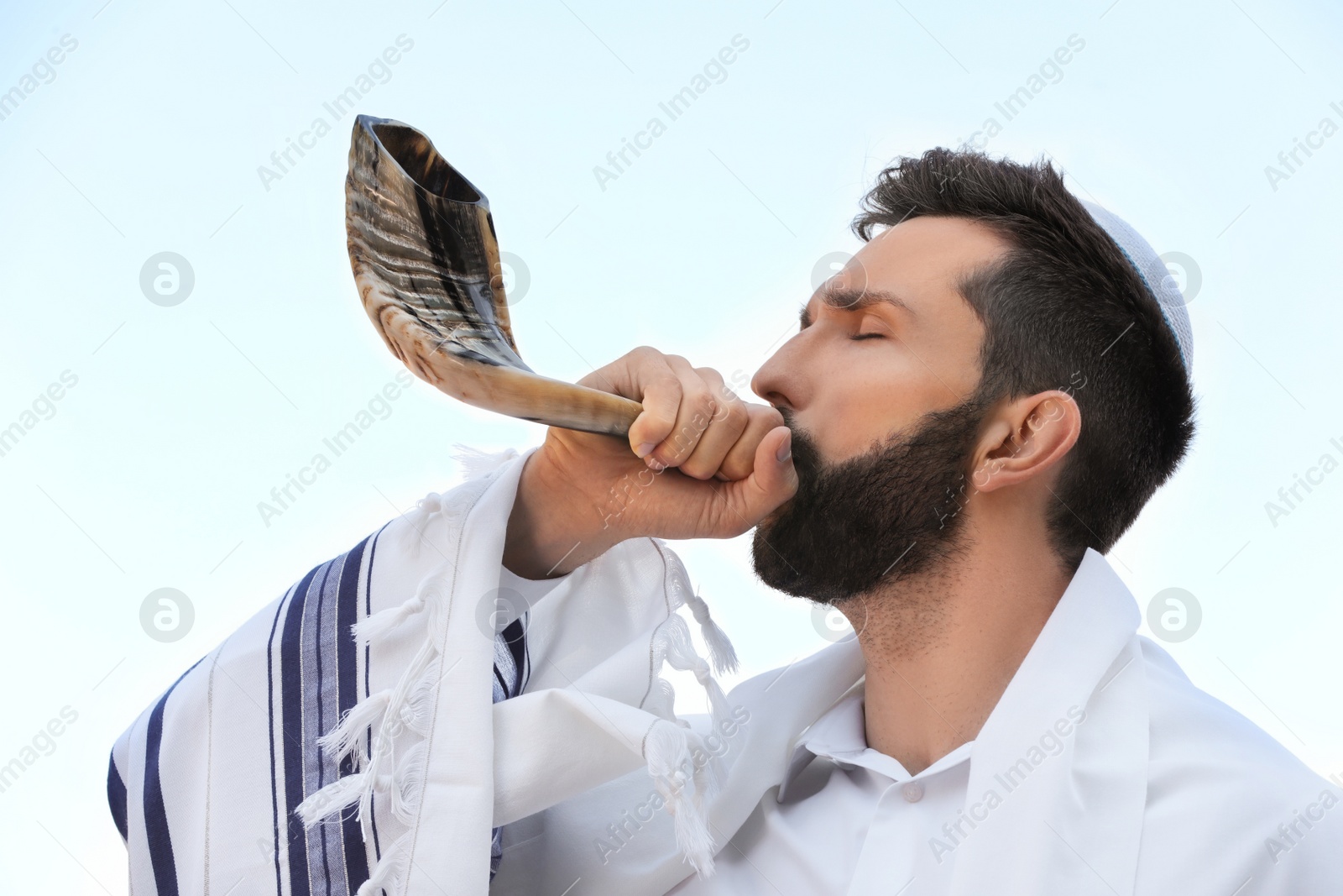 Photo of Jewish man in kippah and tallit blowing shofar outdoors. Rosh Hashanah celebration