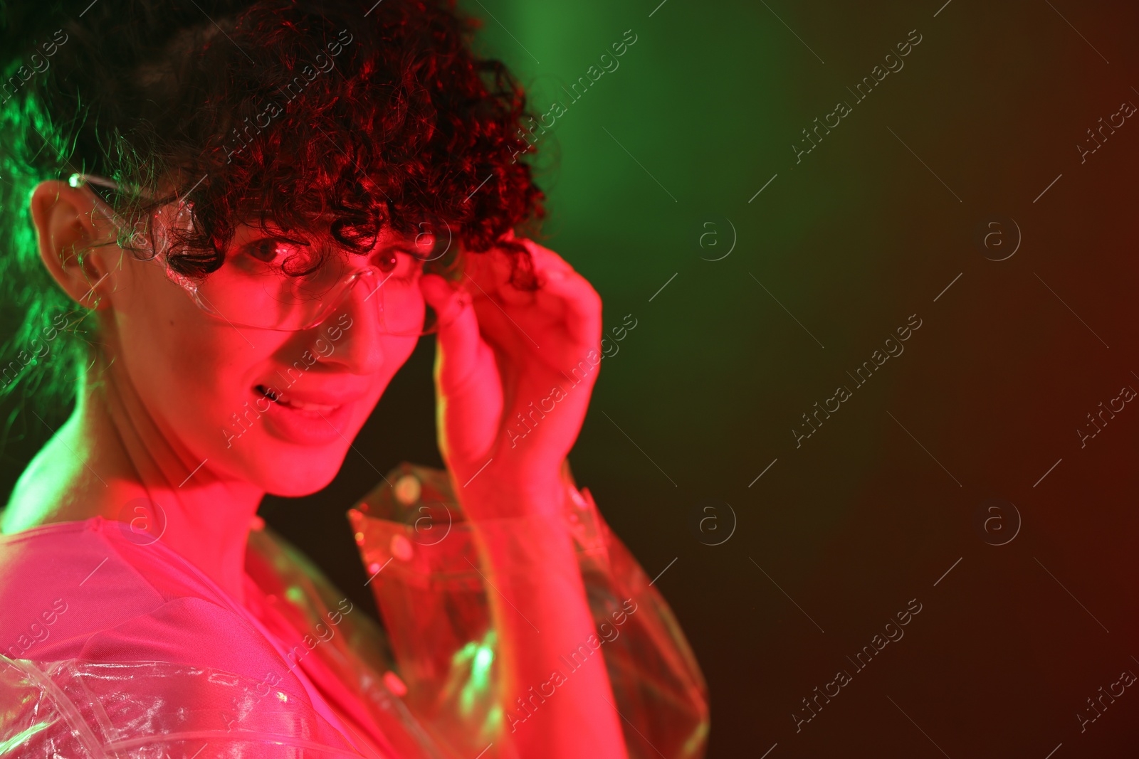 Photo of Beautiful young woman in transparent coat and sunglasses posing on color background in neon lights. Space for text