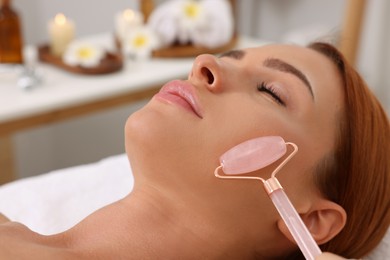 Photo of Young woman receiving facial massage with rose quartz roller in beauty salon, closeup