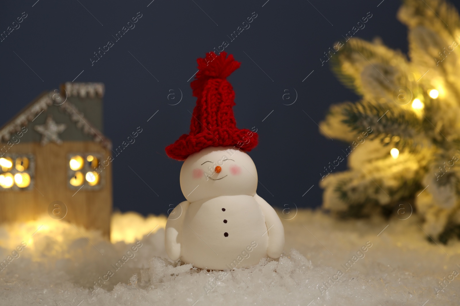 Photo of Cute decorative snowman, hut and Christmas tree on artificial snow against dark background