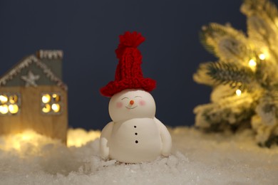 Photo of Cute decorative snowman, hut and Christmas tree on artificial snow against dark background