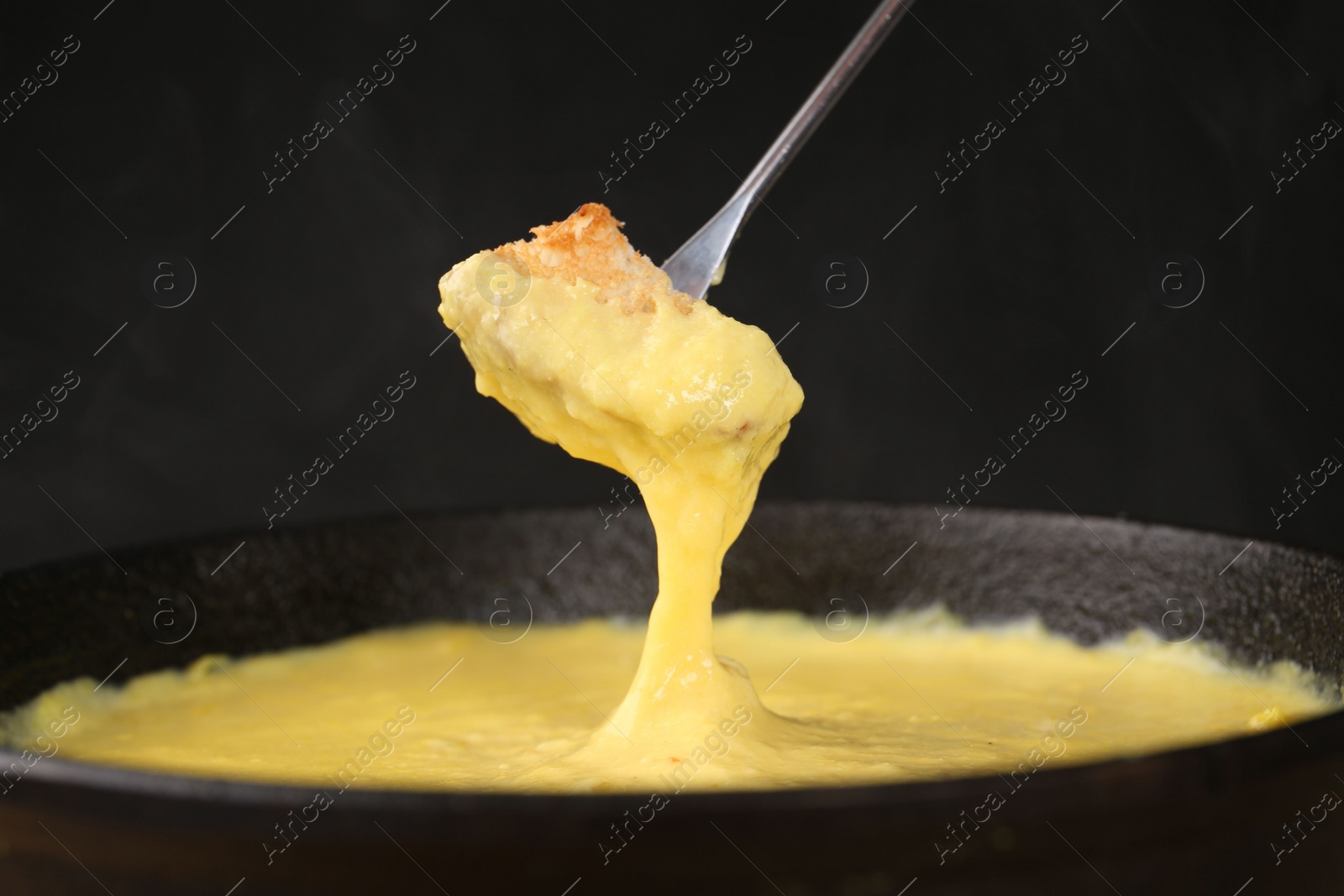 Photo of Dipping piece of bread into fondue pot with tasty melted cheese against dark gray background, closeup