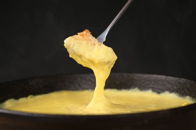 Dipping piece of bread into fondue pot with tasty melted cheese against dark gray background, closeup