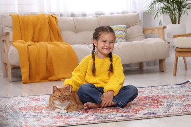 Smiling little girl petting cute ginger cat on carpet at home