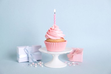 Delicious birthday cupcake with burning candle, sprinkles and gift boxes on light blue background