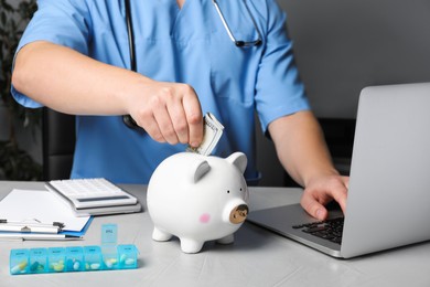 Photo of Doctor putting banknote into piggy bank at table in hospital, closeup. Medical insurance