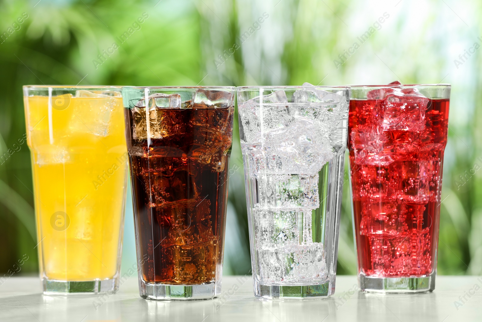 Photo of Glasses of different refreshing soda water with ice cubes on white table outdoors