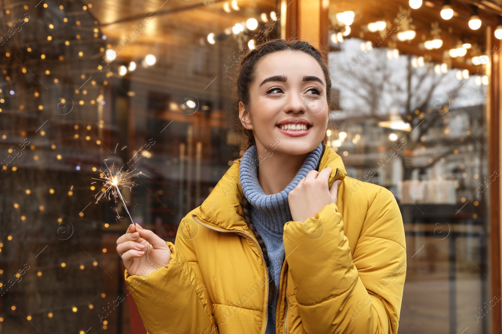 Photo of Young woman with burning sparkler near cafe decorated for Christmas