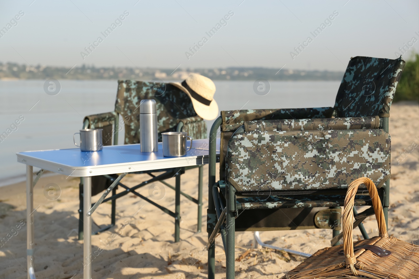 Photo of Camouflage fishing chairs, picnic basket and table on sandy beach near river