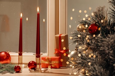Photo of Burning candles, gift boxes and festive decor on window sill near Christmas tree indoors