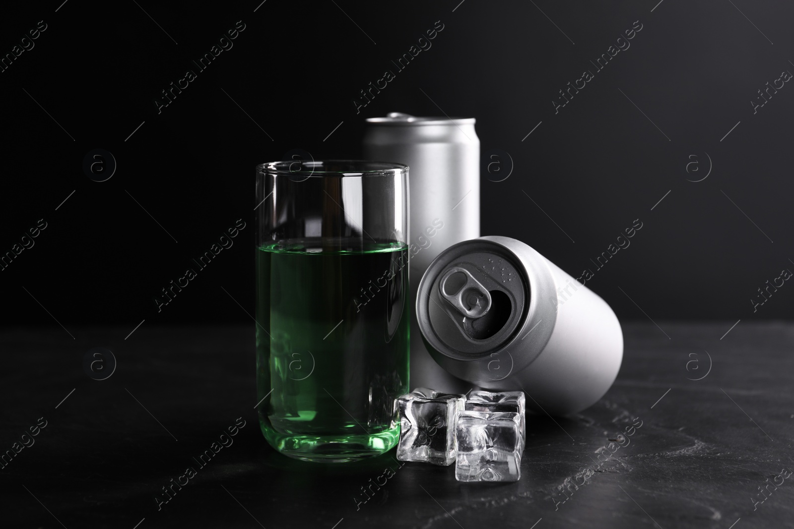 Photo of Energy drink in glass, aluminium cans and ice cubes on black table