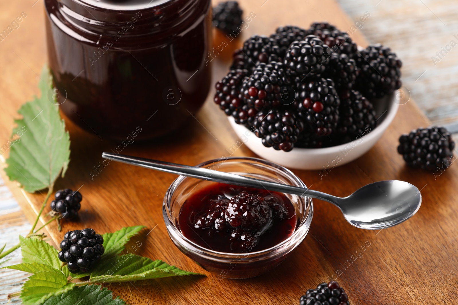 Photo of Fresh ripe blackberries, tasty jam and leaves on wooden board, closeup