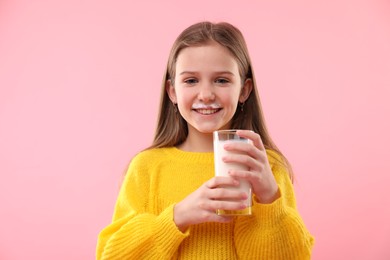 Happy little girl with milk mustache holding glass of tasty dairy drink on pink background