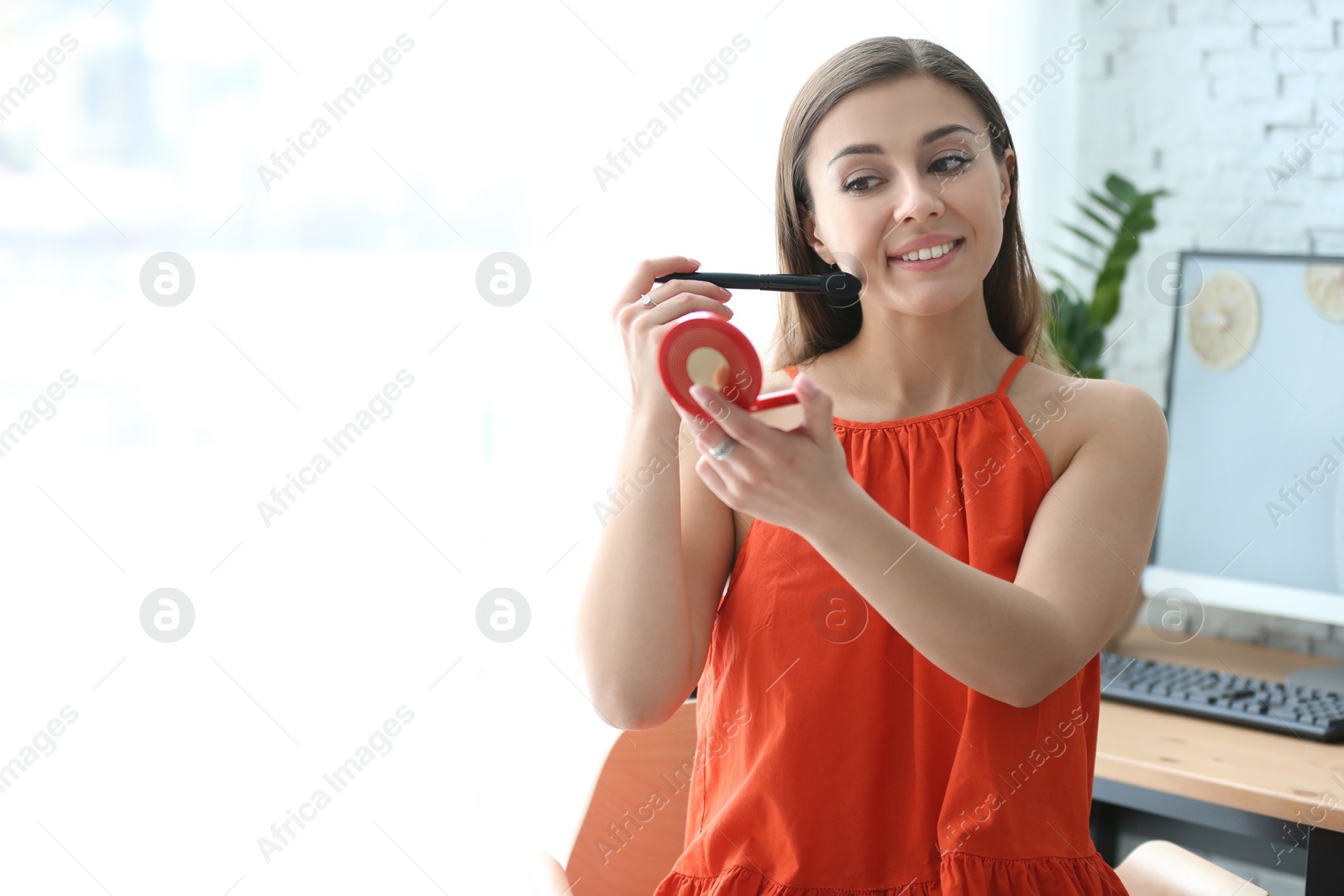 Photo of Young beautiful woman holding mirror while applying makeup in room