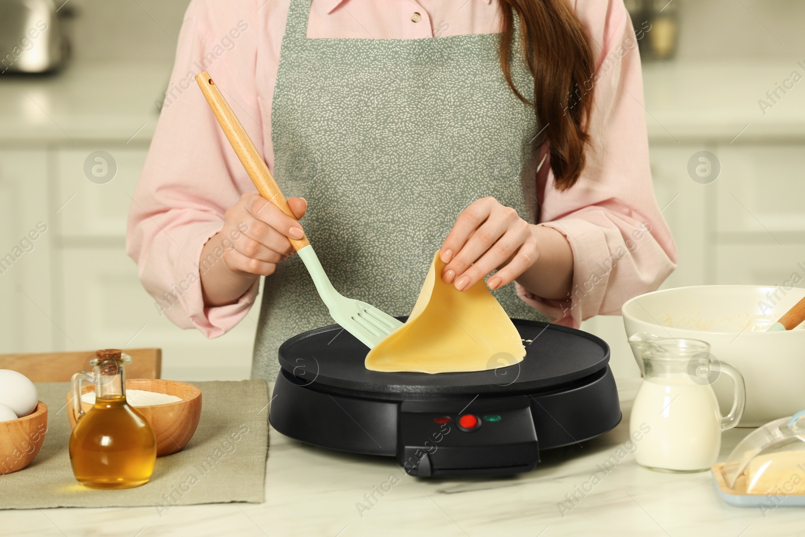 Photo of Woman cooking delicious crepe on electric maker at white marble table in kitchen, closeup