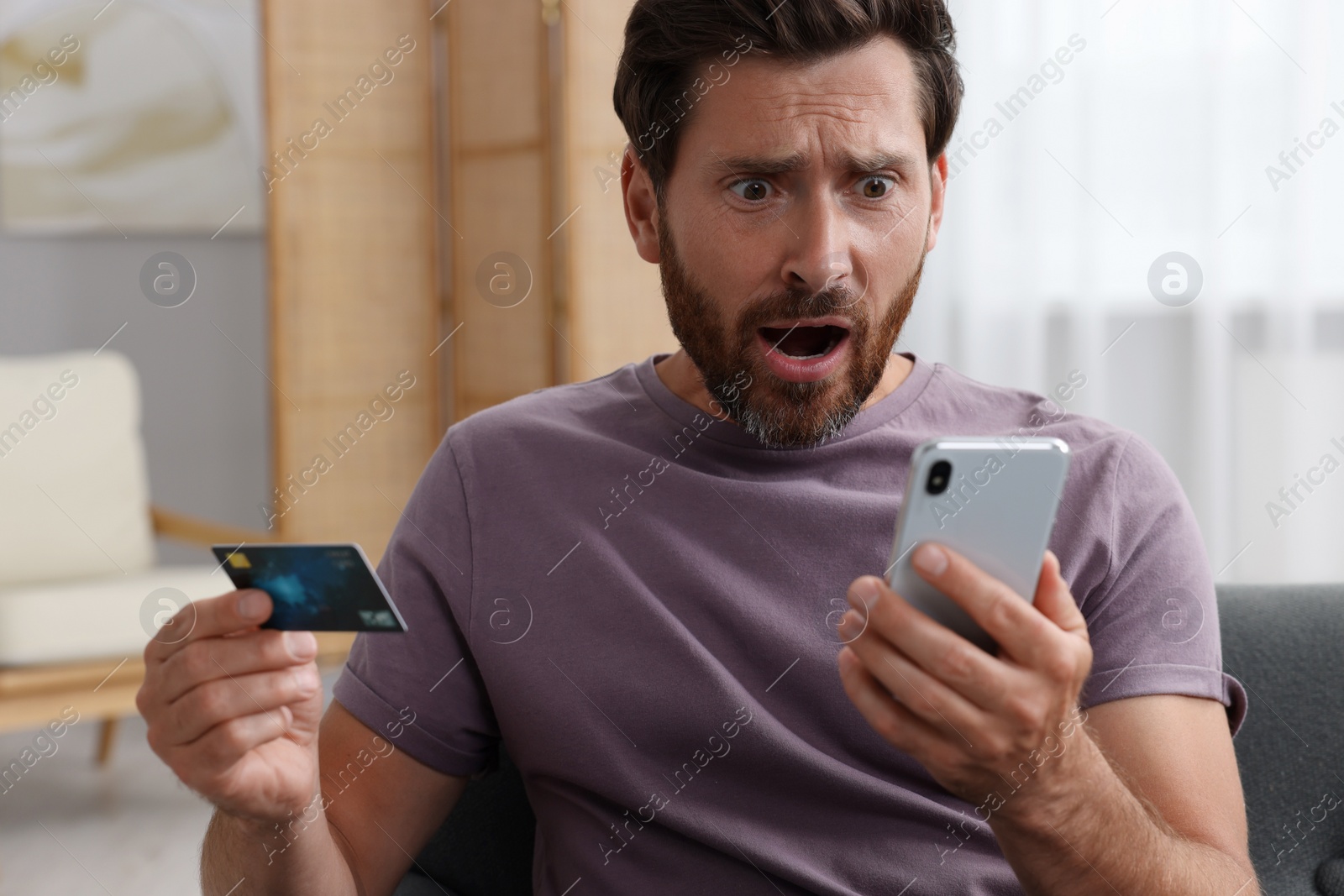 Photo of Shocked man with credit card using smartphone at home. Be careful - fraud