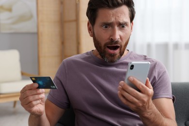 Photo of Shocked man with credit card using smartphone at home. Be careful - fraud