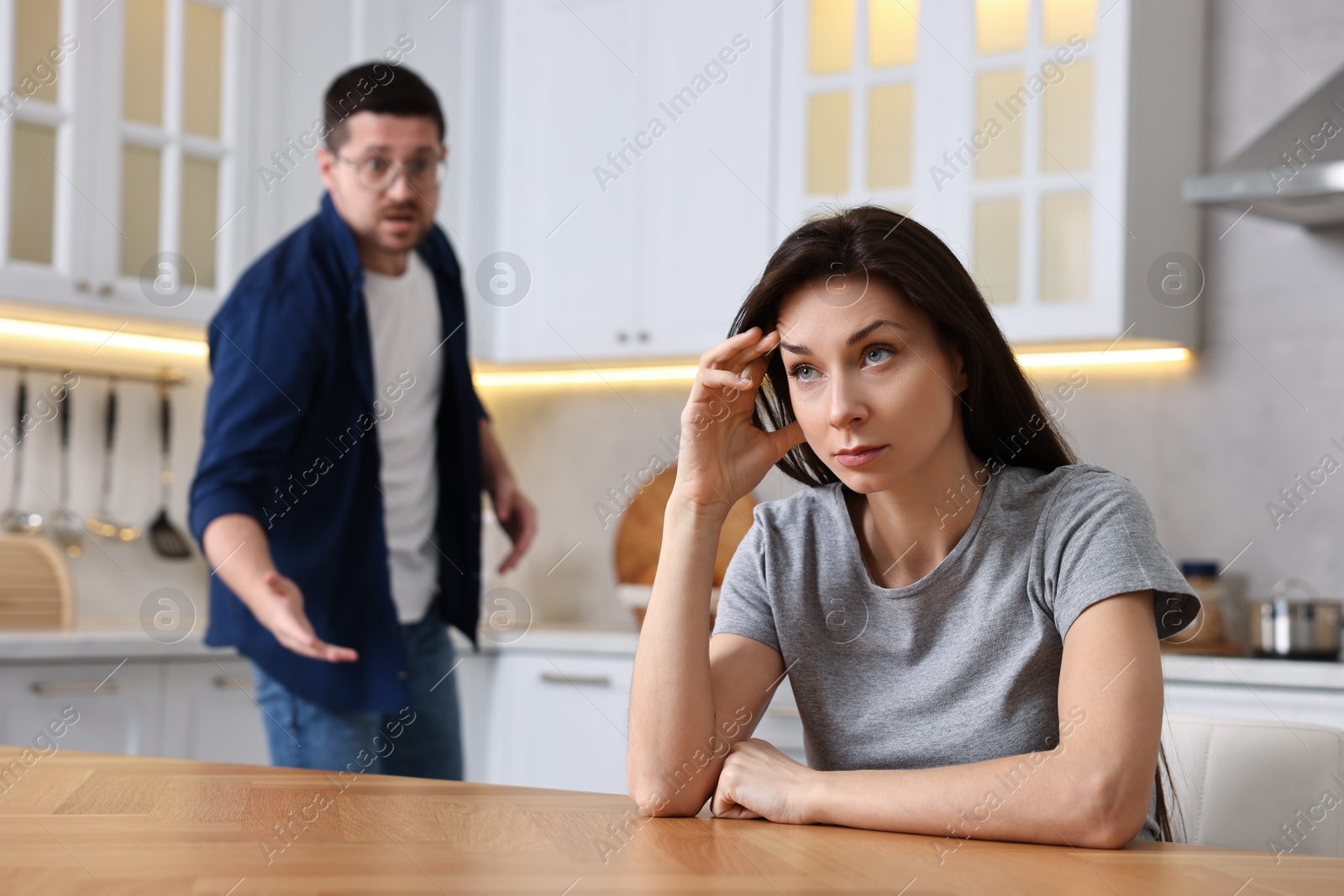 Photo of Stressed wife sitting at table while her angry husband screaming at her in kitchen, selective focus. Relationship problems