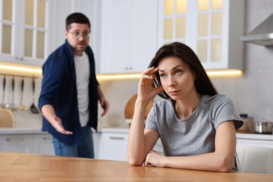 Photo of Stressed wife sitting at table while her angry husband screaming at her in kitchen, selective focus. Relationship problems