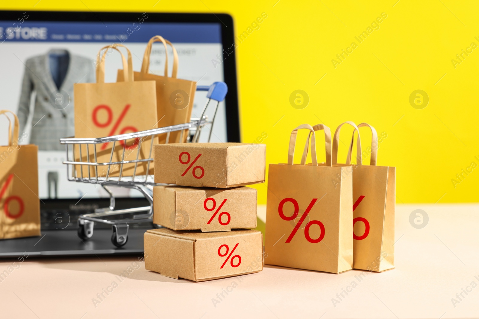 Image of Discount offer. Small shopping bags and boxes with percent signs, mini shopping cart and laptop on beige table