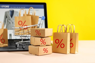 Image of Discount offer. Small shopping bags and boxes with percent signs, mini shopping cart and laptop on beige table