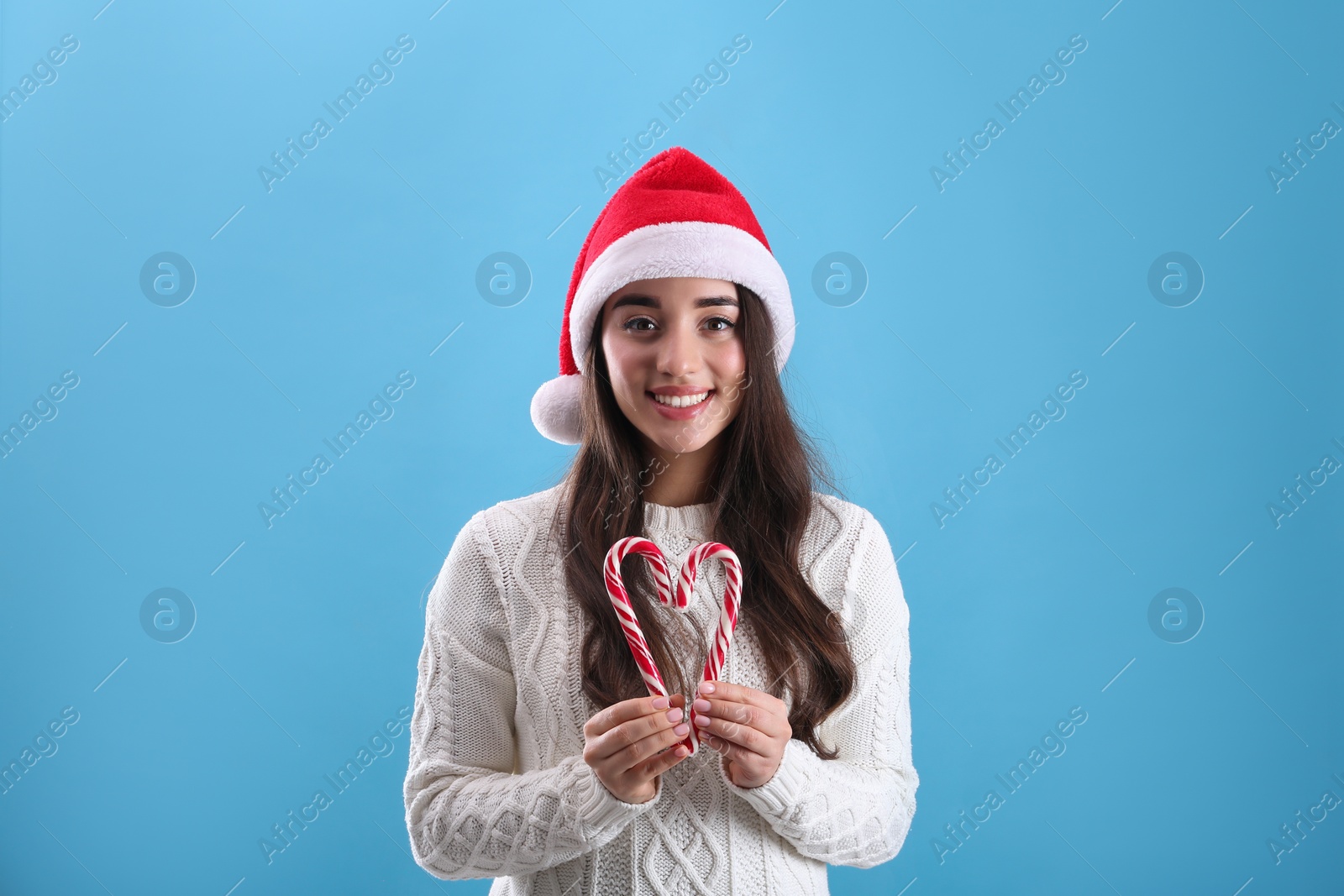 Photo of Beautiful woman in Santa Claus hat making heart with candy canes on light blue background