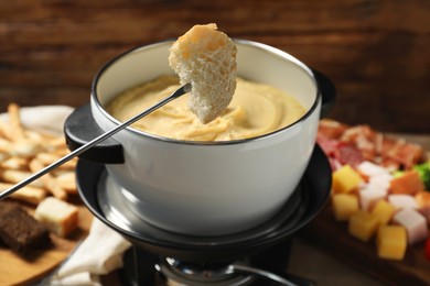 Photo of Fork with piece of bread, melted cheese in fondue pot and snacks on table, closeup