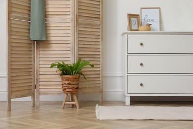 Photo of Stylish room with wooden folding screen and chest of drawers near white wall. Interior design