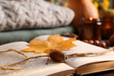 Photo of Book with autumn leaf as bookmark, acorn, scented candles and warm sweaters on wooden table, closeup