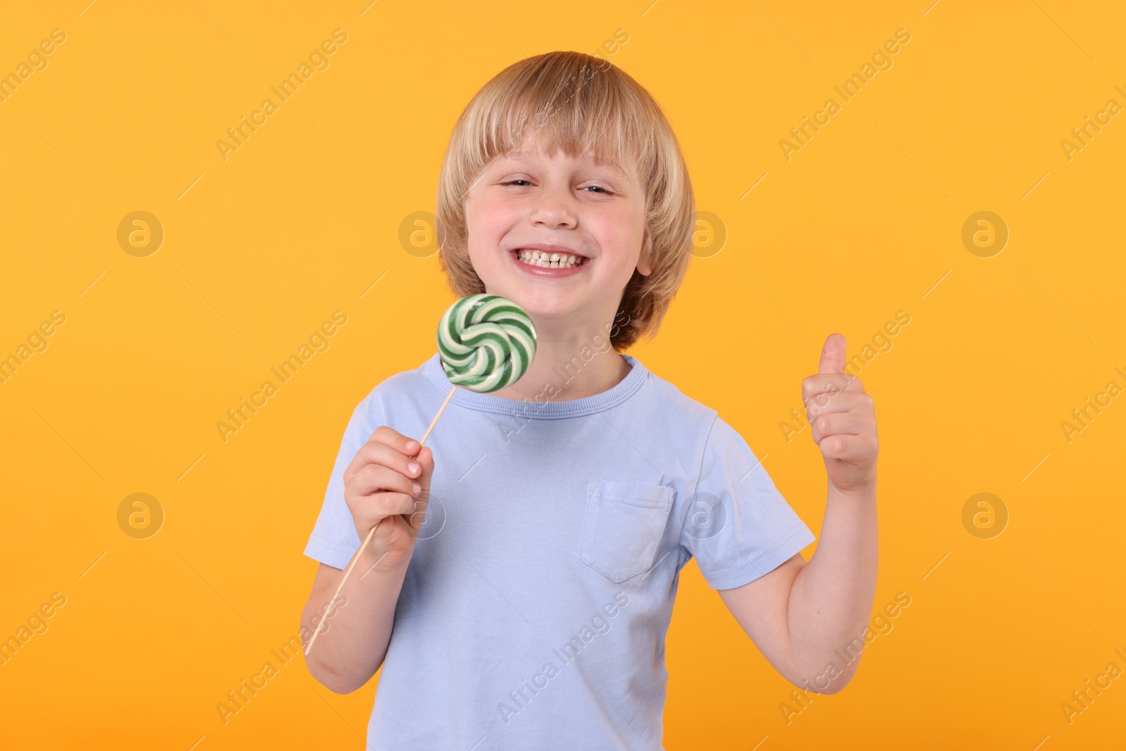 Photo of Happy little boy with bright lollipop swirl showing thumbs up on orange background