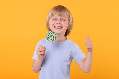 Happy little boy with bright lollipop swirl showing thumbs up on orange background
