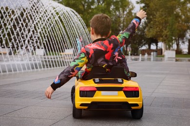Photo of Cute little boy driving children's car near fountain on city street, back view