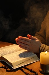 Photo of Woman praying at table with burning candle and Bible, closeup