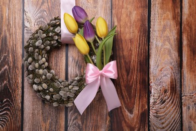 Photo of Wreath made of beautiful willow, tulip flowers and pink bow hanging on wooden background. Space for text