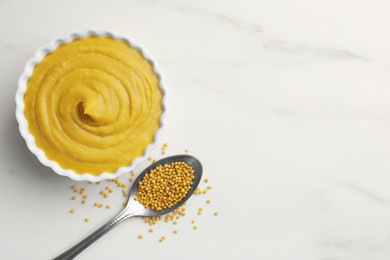 Photo of Tasty mustard sauce in bowl and spoon with dry seeds on white marble table, flat lay. Space for text