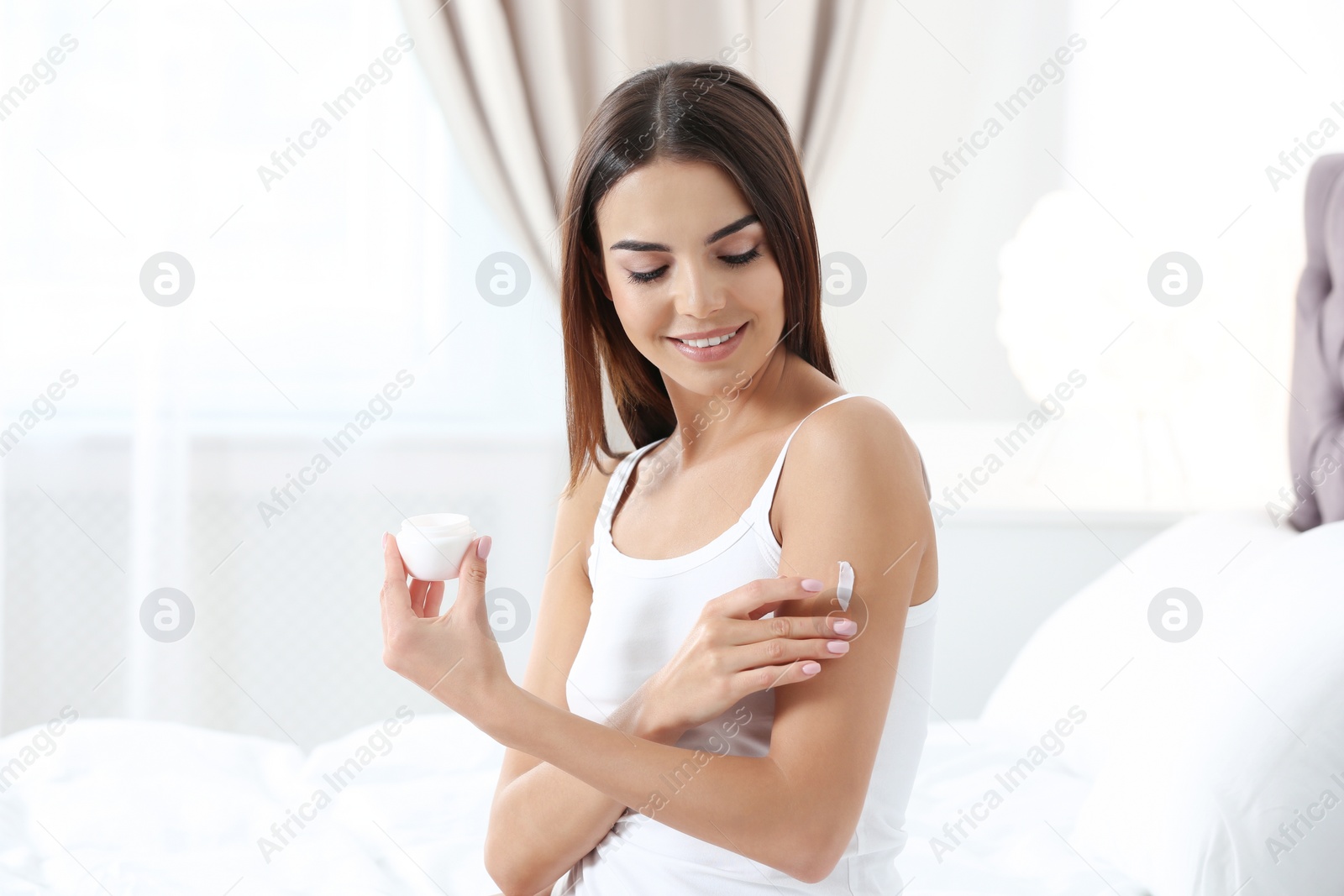 Photo of Young woman with perfect smooth skin applying cream in bedroom. Beauty and body care