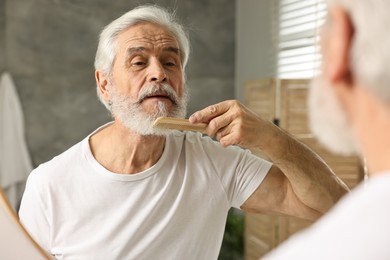 Photo of Senior man with mustache combing beard near mirror in bathroom