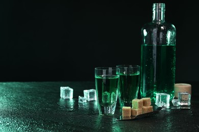 Photo of Absinthe in shot glasses, spoon, brown sugar and ice cubes on gray table against dark background, space for text. Alcoholic drink