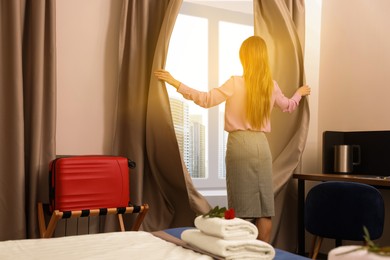 Young woman opening window curtains in hotel room, back view
