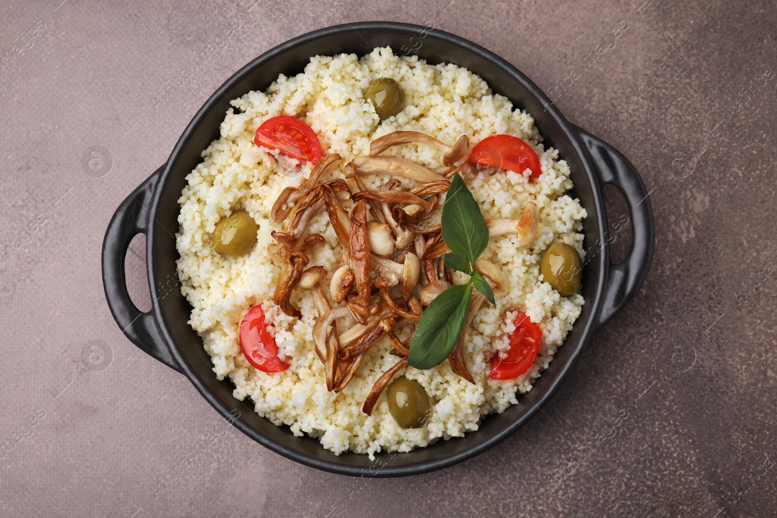 Photo of Frying pan of tasty couscous with mushrooms, olives and tomatoes on brown textured table, top view