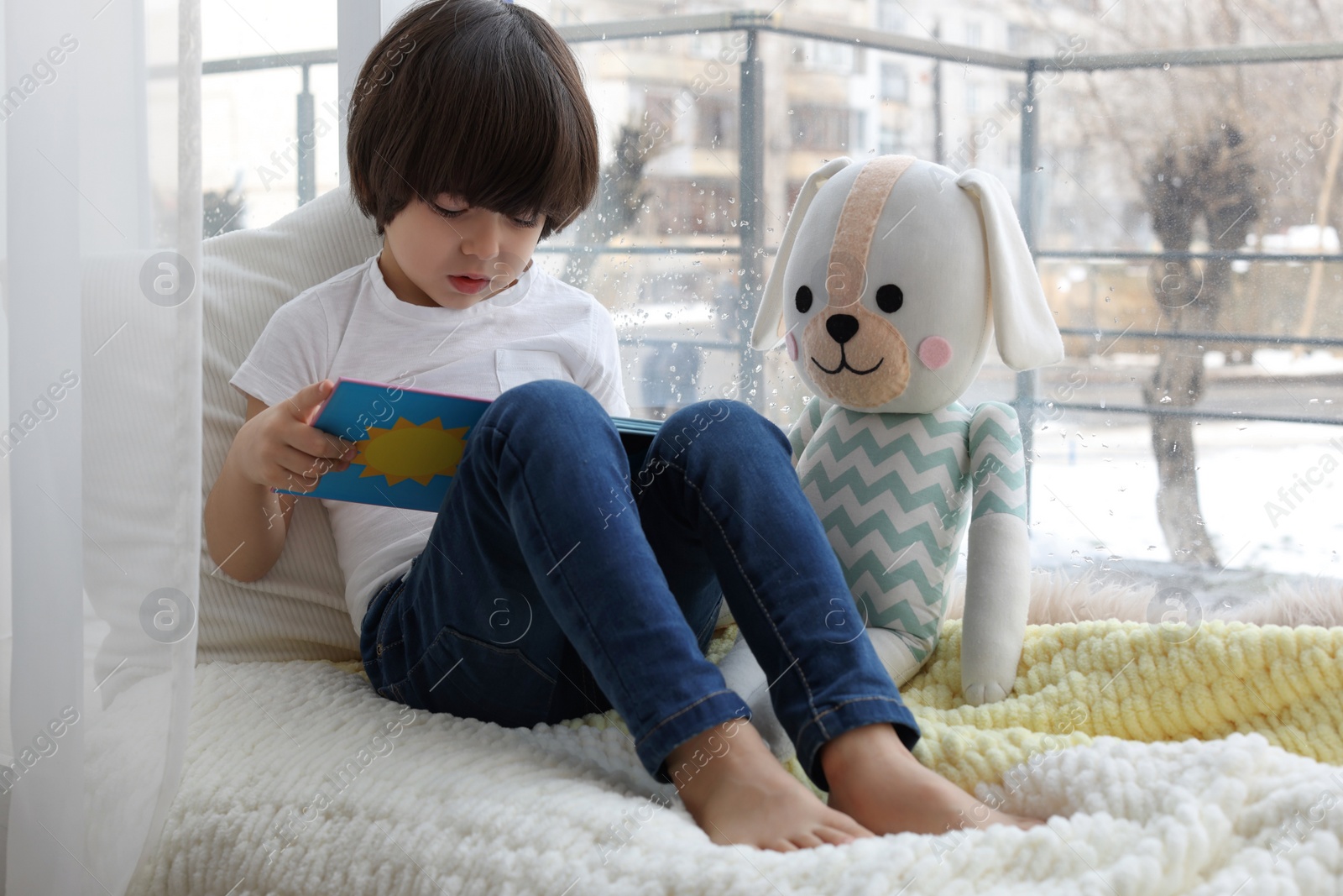 Photo of Cute little boy with toy reading book near window at home
