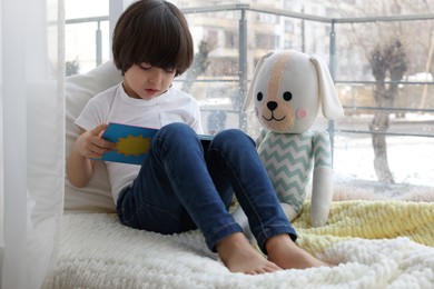 Cute little boy with toy reading book near window at home