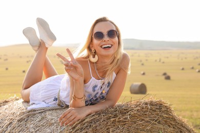 Photo of Beautiful hippie woman showing peace sign on hay bale in field, space for text