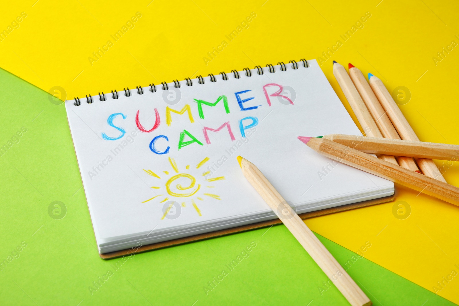 Photo of Notebook with written text SUMMER CAMP and different pencils on color background